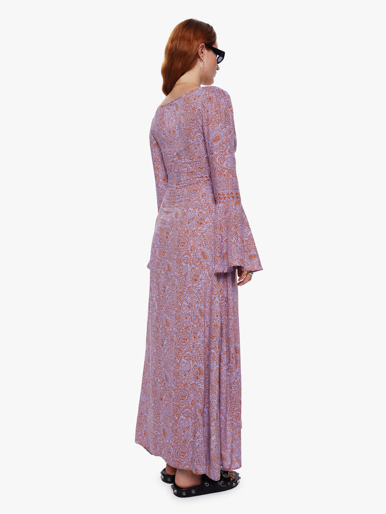 A back view of a woman wearing an orchid-purple and rust floral print, full length dress with bell sleeves and keyhole neckline.