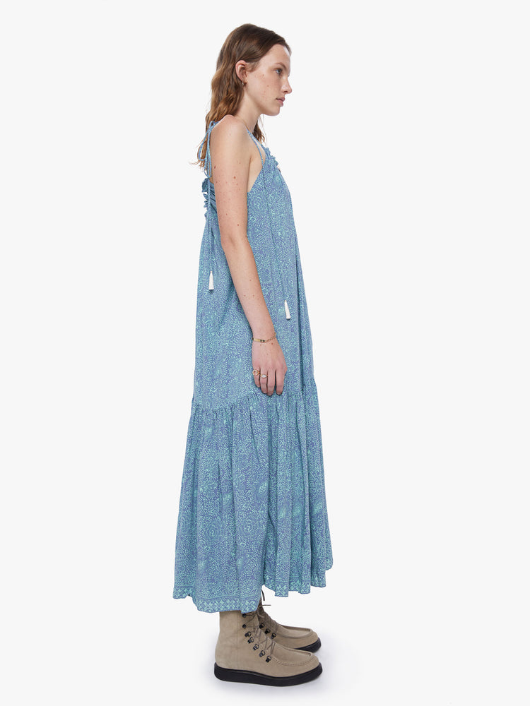 Side view of a women wearing a periwinkle delicate floral print with a square neckline, slim adjustable straps that tie and a floaty tiered skirt just above the ankle