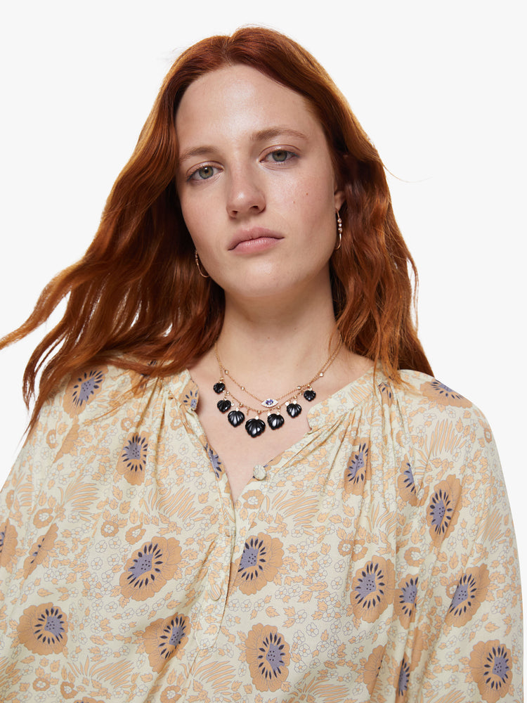 Close up view of a woman wearing a sand colored floral print v-shaped neckline top with covered buttons and subtle pleats below the collar emphasize loose feel
