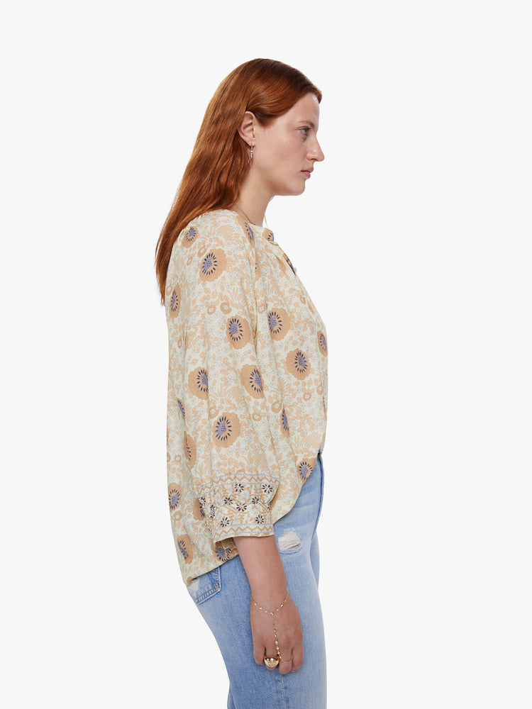 Side view of a woman wearing a sand colored floral print v-shaped neckline top with covered buttons and subtle pleats below the collar emphasize loose feel
