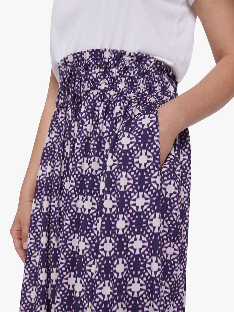 Side close up view of a womens mid length skirt featuring a purple and white print, a rouched high waist, concealed side pockets, and a flowy fit.
