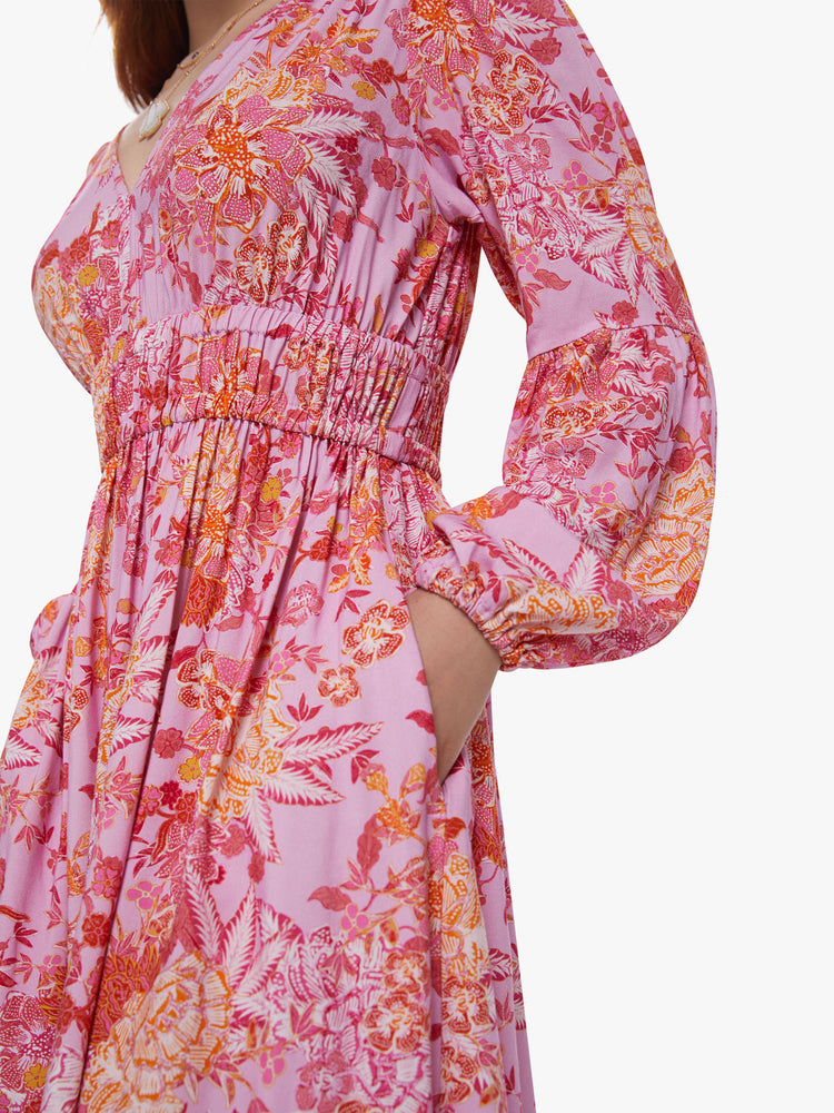 Side close up view of a womens midi pink and orange floral dress, featuring long billow sleeves, side hidden pockets, a rouched waist and v neck.
