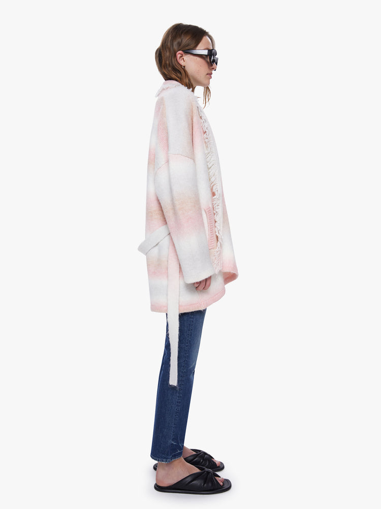 Side view of a womens long cardigan, featuring soft pink and white stripes, dropped sleeves, fringe trim, and a belted tie.