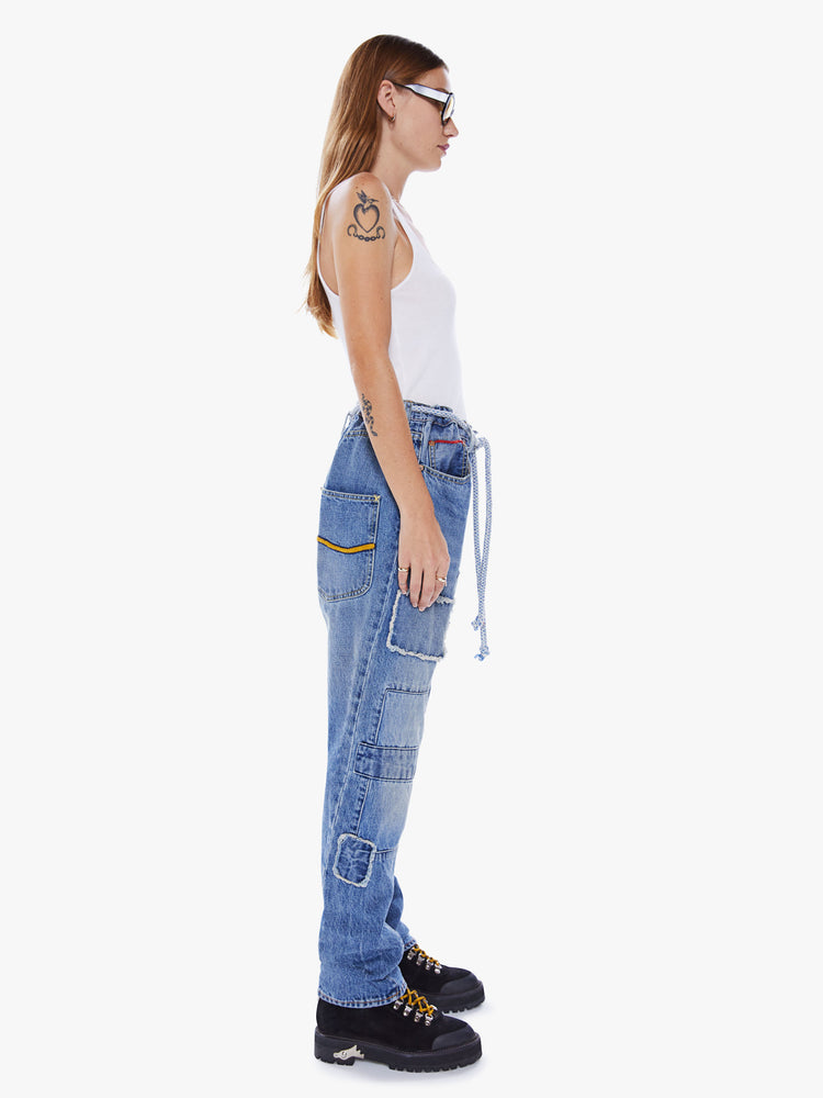 A side view of a woman wearing a medium blue denim pant with patchwork repairs and a rope belt