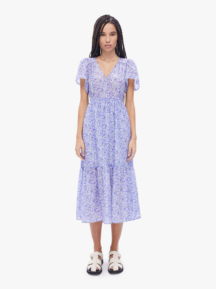 Front full body view of a woman in a midi dress from Xirena, made from a blend of cotton and silk in periwinkle floral print the dress is designed with a Vneck, sheer short sleeves, an elastic high waist and a calf length skirt with flowy gathered tiers