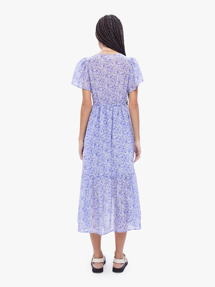 Back full body view of a woman in a midi dress from Xirena, made from a blend of cotton and silk in periwinkle floral print the dress is designed with a Vneck, sheer short sleeves, an elastic high waist and a calf length skirt with flowy gathered tiers