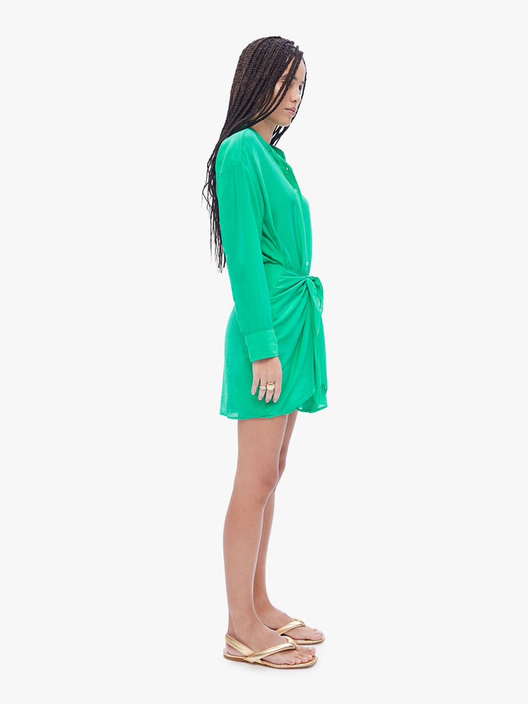 Side full body view of a woman in Xirena long sleeve mini dress made from a blend of cotton and silk in a bright green hue, and features a vneck, buttons down the front and a layered, gathered skirt that ties at the waist.