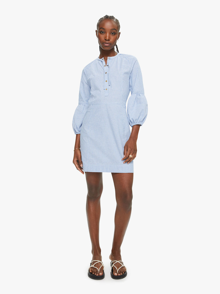 Front view of Vita dress features a buttoned V-neck, 3/4-length sleeves with gathered elastic hems, and a narrow fit. Made of 100% cotton in a light blue chambray with subtle white stripes, the slim-fitting dress is soft and lightweight.