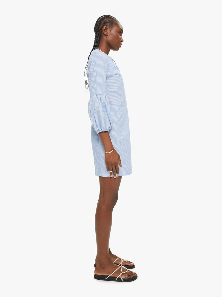 Side view of Vita dress features a buttoned V-neck, 3/4-length sleeves with gathered elastic hems, and a narrow fit. Made of 100% cotton in a light blue chambray with subtle white stripes, the slim-fitting dress is soft and lightweight.