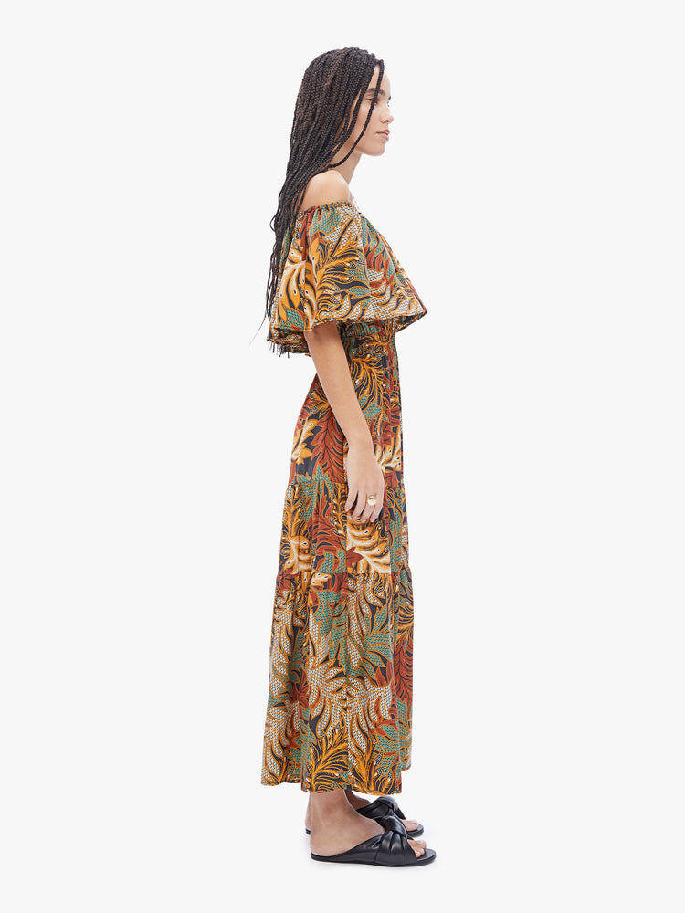 Side full body view of a woman in a Naomi dress by Natalie Martin made from 100% silk in a brown, green, burnt orange and cream leaf print. Maxi dress features an off the shoulder elastic neckline with an oversized ruffle, a fitted waist and a tiered ankle-length skirt