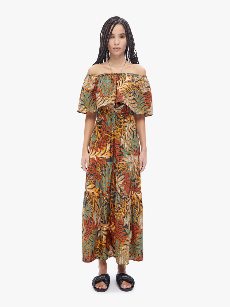 Front full body view of a woman in a Naomi dress by Natalie Martin made from 100% silk in a brown, green, burnt orange and cream leaf print. Maxi dress features an off the shoulder elastic neckline with an oversized ruffle, a fitted waist and a tiered ankle-length skirt
