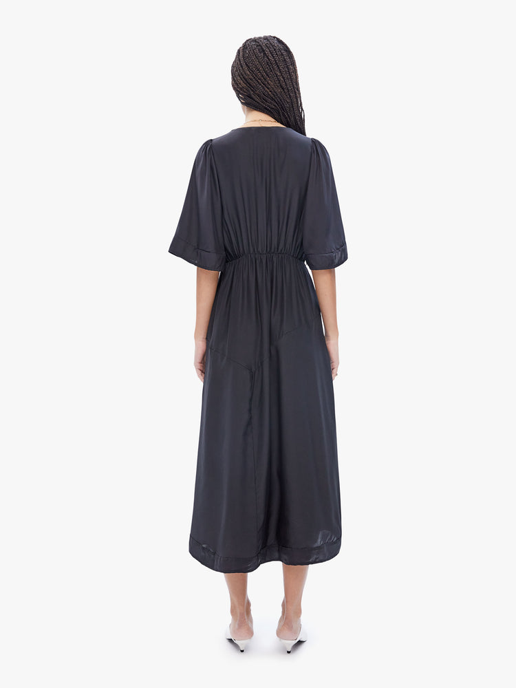 Back full body view of a woman in maxi dress by Natalie Martin, made from 100% silk in black and features a wrapped v-neck, 3/4 length balloon sleeves, an elastic waist and an asymmetrical hem