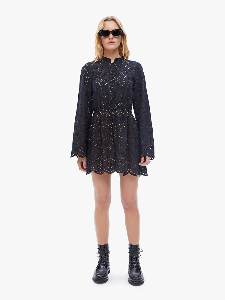 Front full body view of a woman in a Cleo dress from Natalie Martin, made from a semi-sheer 100% cotton in a rich black hue with openwork details, the mini dress features a crewneck with a short collar, long flowy sleeves, a fitted waist and zig-zag hems