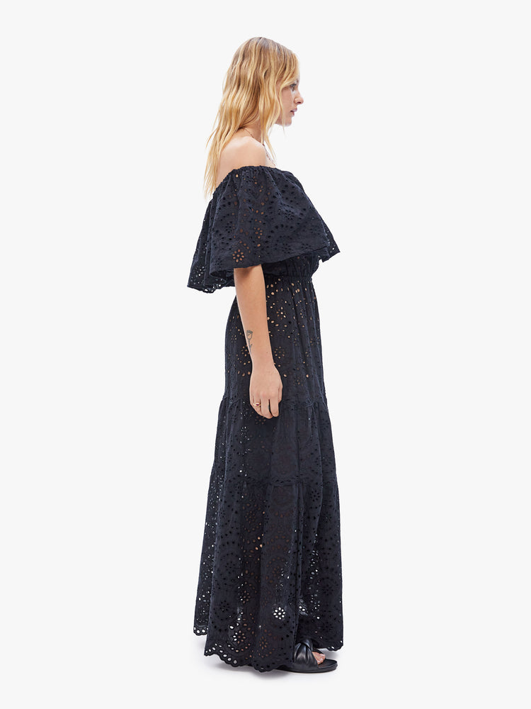 Side full body view of a woman in a Naomi dress crafted by Natalie Martin, made from semi sheer 100% cotton in a rich black hue with openwork details features an off the shoulder elastic neckline with an oversized ruffle, a fitted waist and a tiered ankle-length skirt
