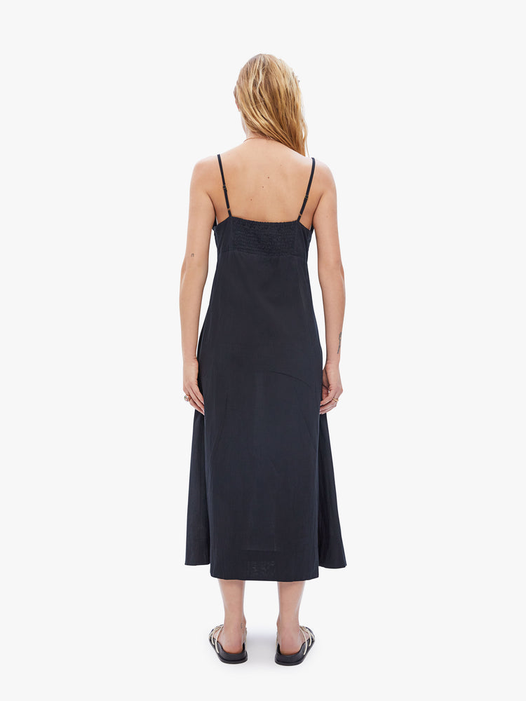 Back full body view of a woman in a Sophie dress created by Natalie Martin in a 100% cotton in a rich black hue with slim staps, a v-neck with a bow at the chest, an ankle-length hem and a flowy a line fit