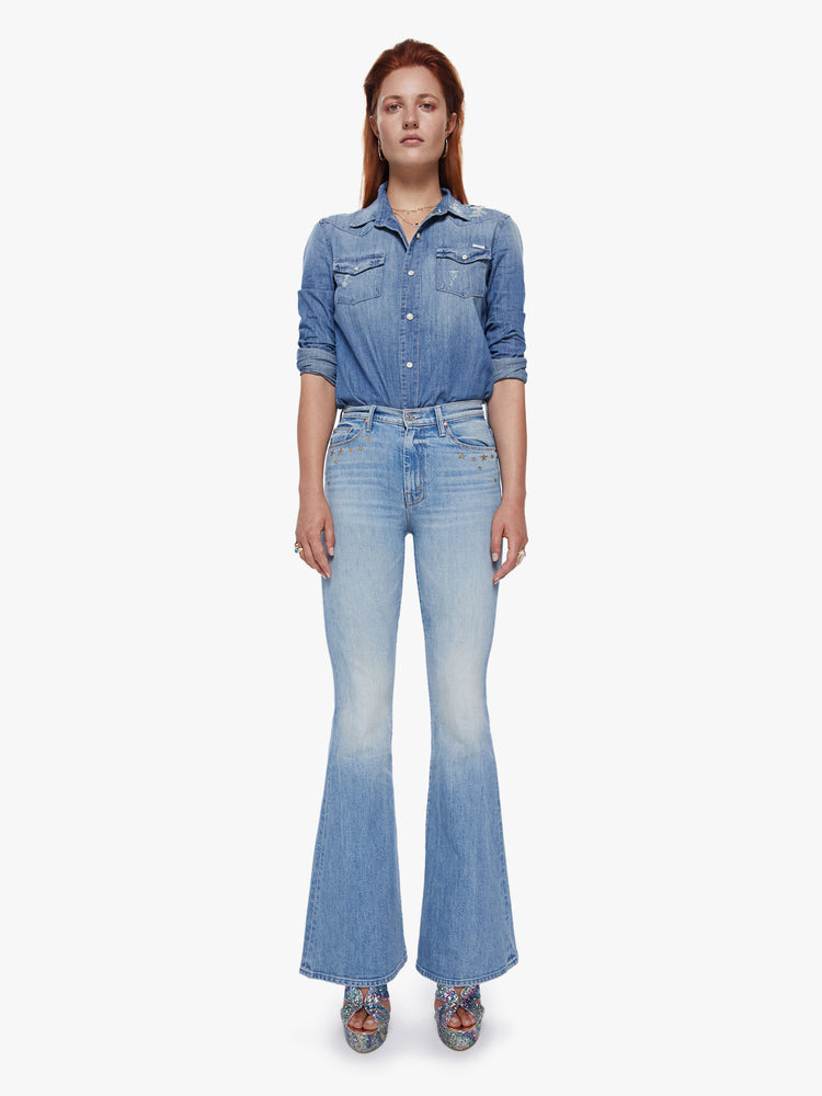 Front full body view of a woman in a high rise flare jean with a long 34.5 inch inseam and a clean hem cut from semi-rigid denim in a light blue wash with whiskering, fading and glittery gold stars and text across the back