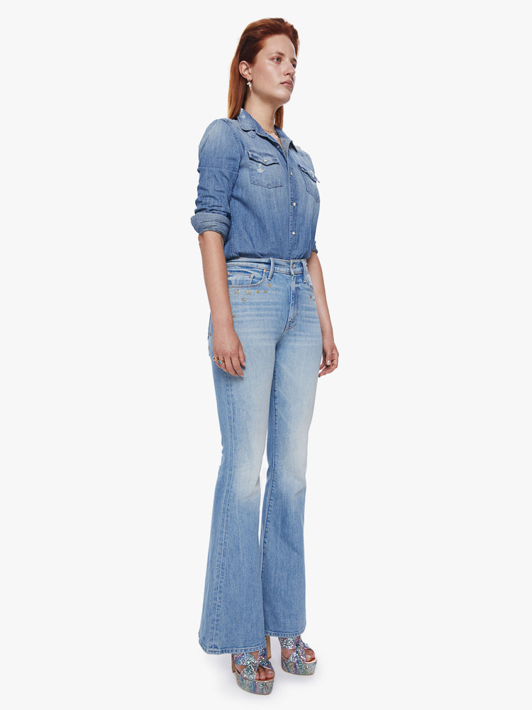 Side angle full body view of a woman in a high rise flare jean with a long 34.5 inch inseam and a clean hem cut from semi-rigid denim in a light blue wash with whiskering, fading and glittery gold stars and text across the back