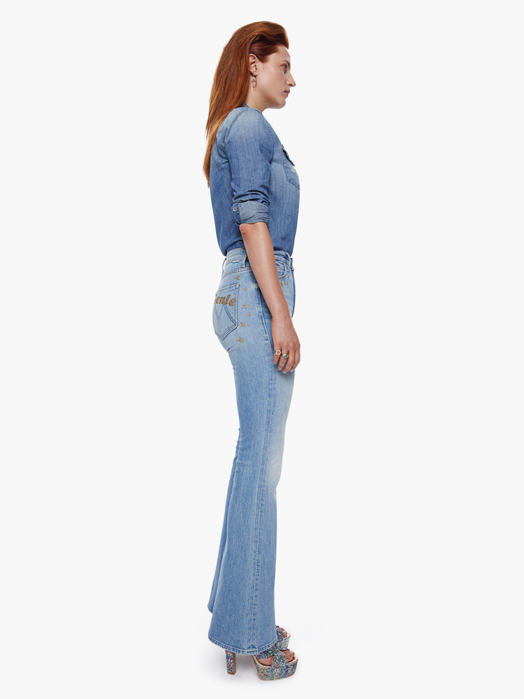 Side full body view of a woman in a high rise flare jean with a long 34.5 inch inseam and a clean hem cut from semi-rigid denim in a light blue wash with whiskering, fading and glittery gold stars and text across the back