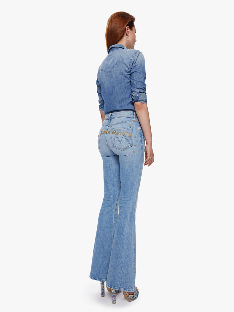 Back full body view of a woman in a high rise flare jean with a long 34.5 inch inseam and a clean hem cut from semi-rigid denim in a light blue wash with whiskering, fading and glittery gold stars and text across the back