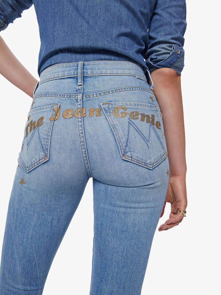 Close up view of a woman in a high rise flare jean with a long 34.5 inch inseam and a clean hem cut from semi-rigid denim in a light blue wash with whiskering, fading and glittery gold stars and text across the back
